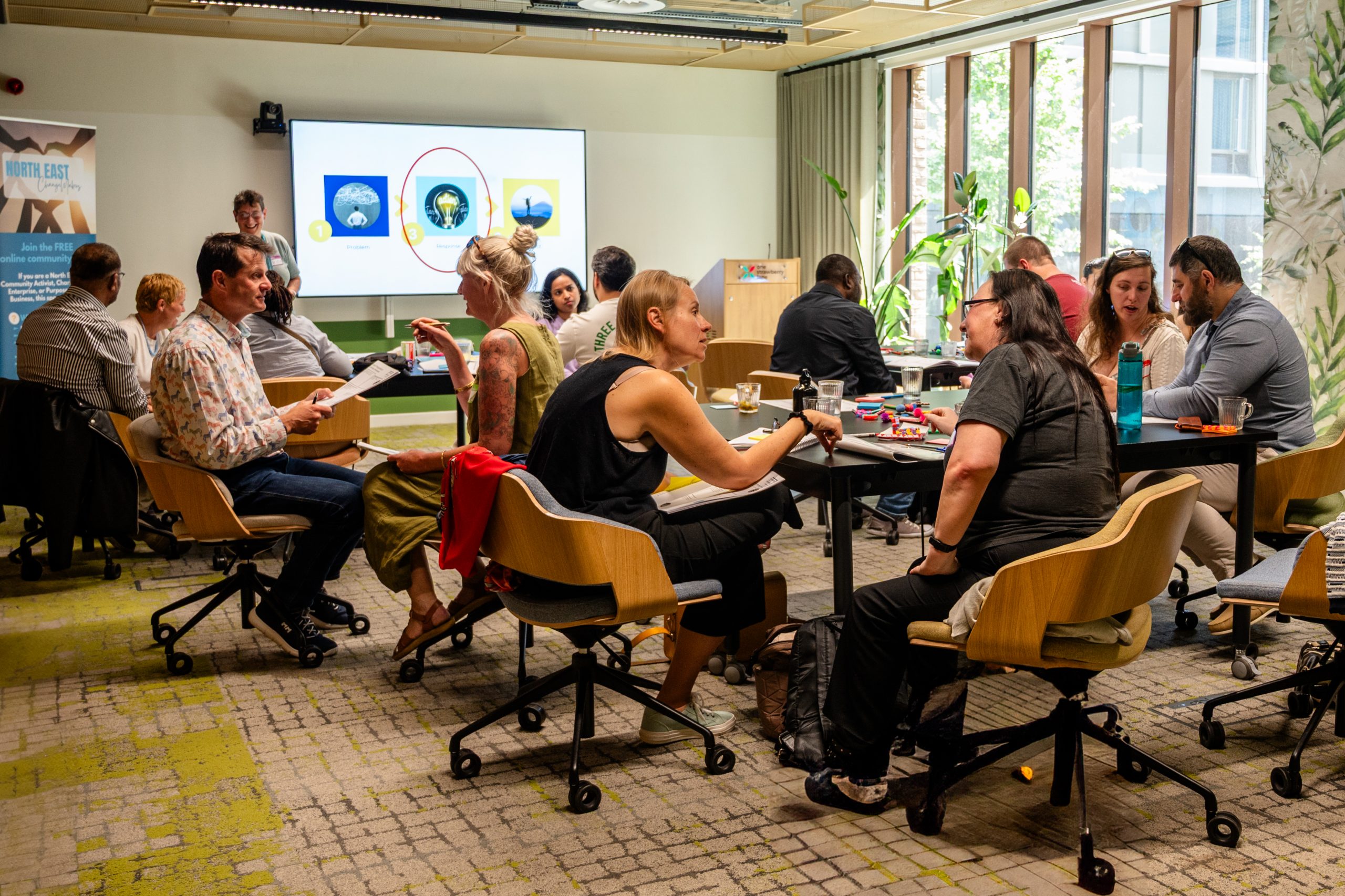 Participants sat around tables chatting. The room is light with big windows.